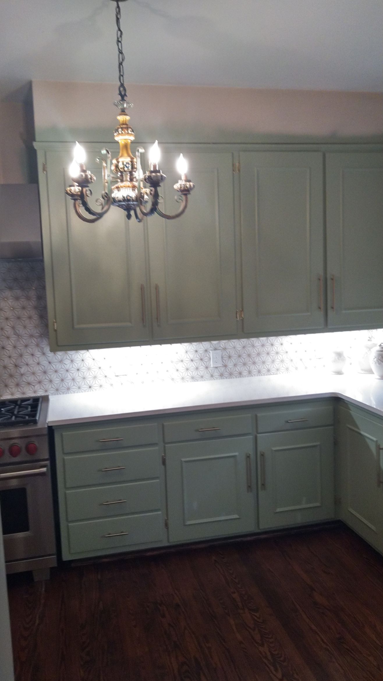 Small kitchen with pale green cabinets, white backsplash, dark wood floor, and a hanging chandelier.