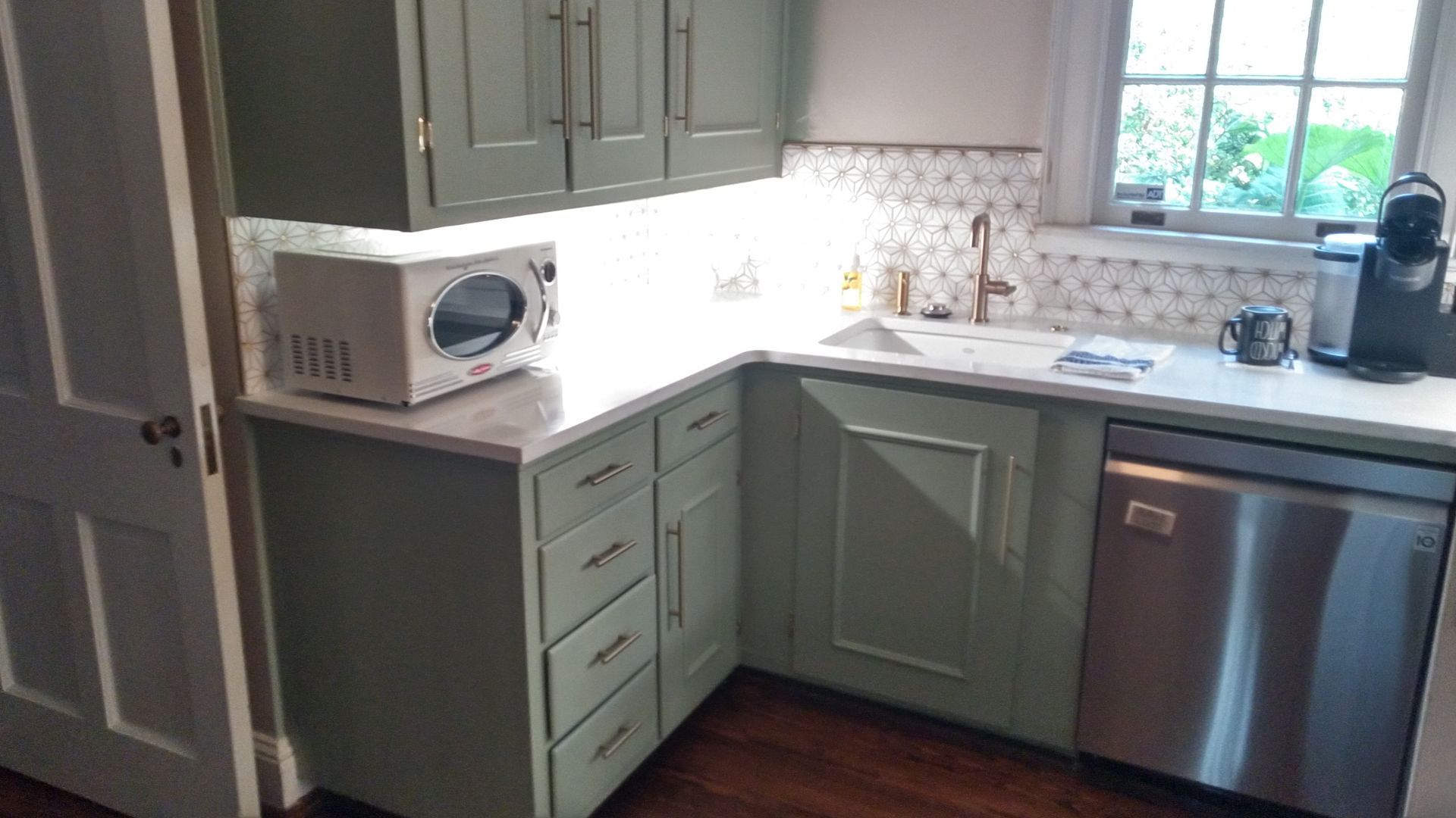 Bright kitchen corner with green cabinets, white countertop, microwave, sink by window, and stainless dishwasher.