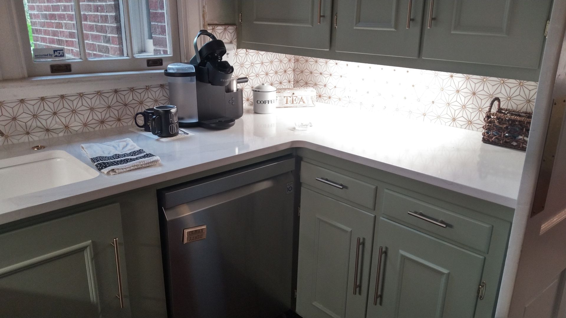 Kitchen counter with white countertop, olive cabinets, sink, coffee maker, and daylight from window.