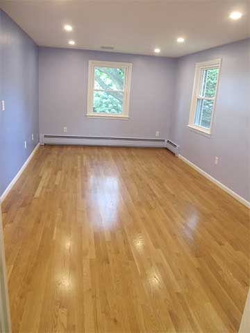 Empty room with light wood floors, pale walls, recessed lights, and two windows.