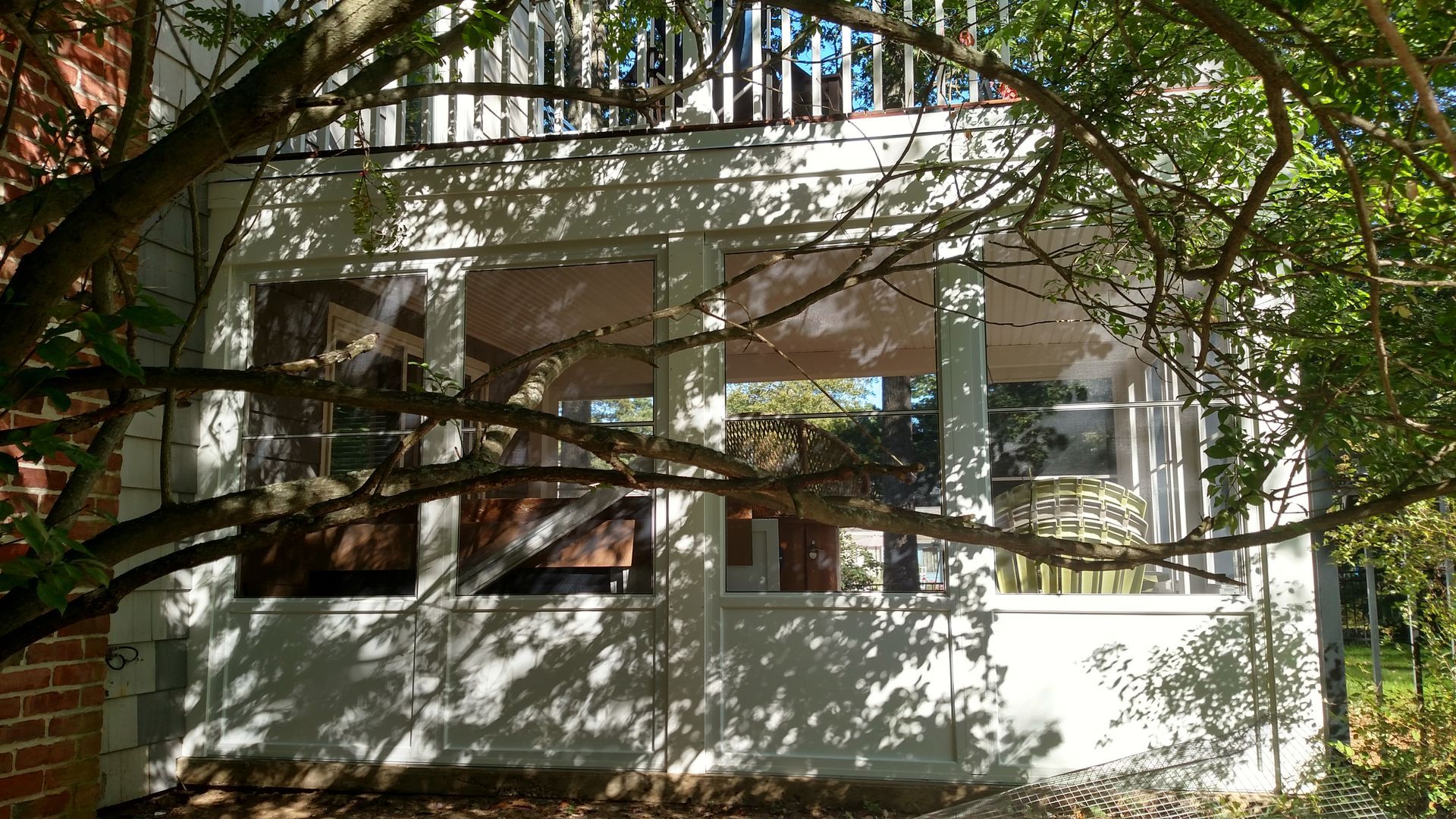 White house porch with large windows, shaded by tree branches and surrounded by greenery.