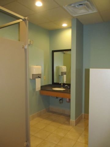 Public restroom stall with sink, mirror, and paper towel dispenser on light blue walls and beige tile floor