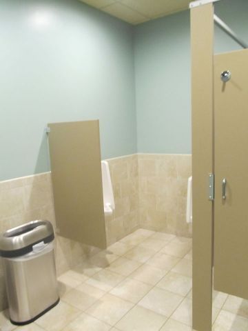 Public restroom stall area with beige tiled walls, a trash can, and open stall doors