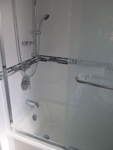 White tiled shower with chrome grab bar, handheld showerhead, and glass door.