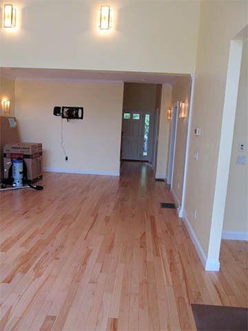 Empty living room with light wood floors, beige walls, and wall lights, viewed toward a hallway.