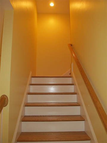 Narrow indoor staircase with white steps, wooden handrail, and warm yellow lighting