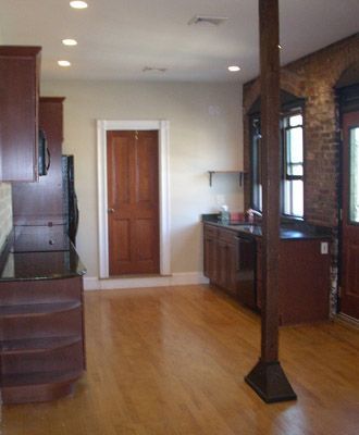 Modern kitchen with wood cabinets, hardwood floor, and a brick-accent wall beside a dining area.
