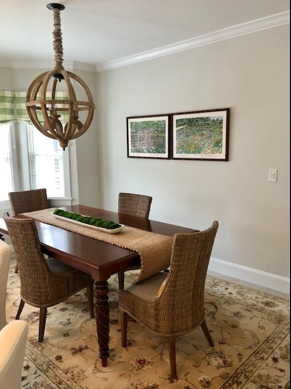 Dining room with wicker chairs, wooden table, pendant light, and framed wall art on a patterned rug.
