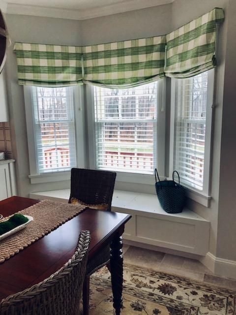 Dining nook with bay windows, green plaid valance, wooden table, chair, rug, and a black bag on the bench.