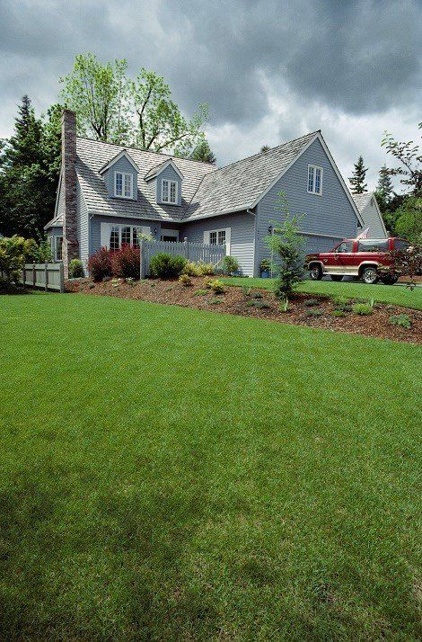 Blue-gray house with dormer windows and a red truck beside a lush green lawn under cloudy skies