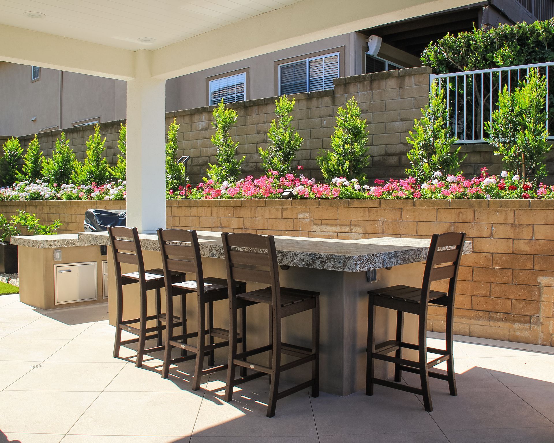 Outdoor patio with a stone dining table, bar stools, and a raised flower bed along a brick wall.