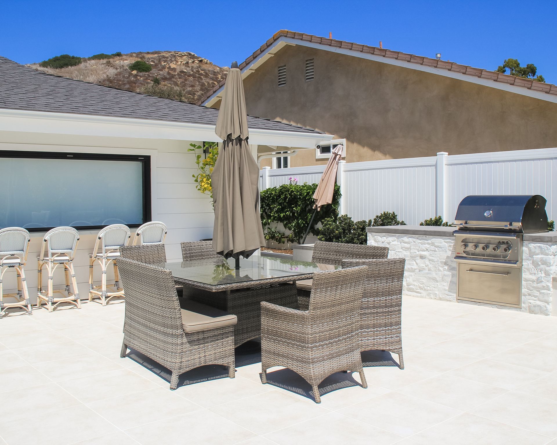Patio with wicker dining set, closed umbrella, lounge chairs, and grill beside a house and white fence