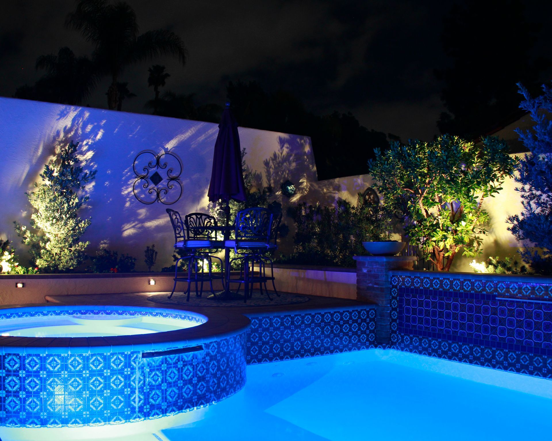 Blue-lit backyard pool at night with tiled spa, lounge chairs, and plants against a white wall
