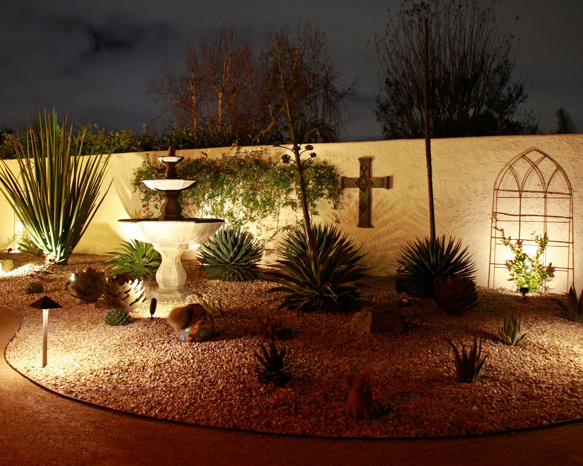 Illuminated desert garden with cacti, rocks, a cross on a stucco wall, and a decorative arch at night