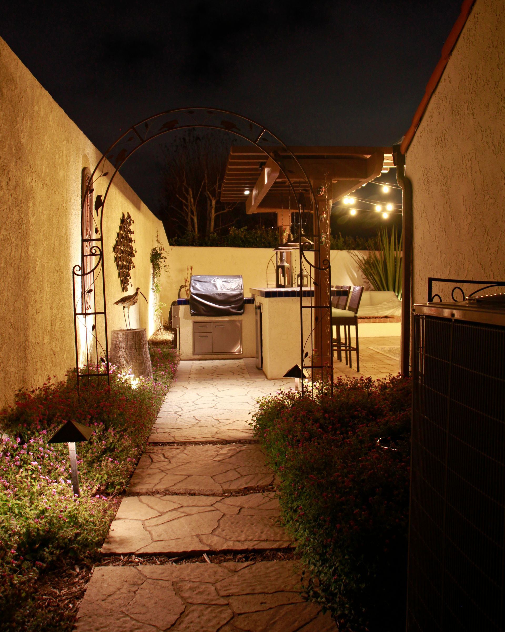 Narrow garden walkway at night, lit by warm string lights, leading to a patio with chairs and a grill.