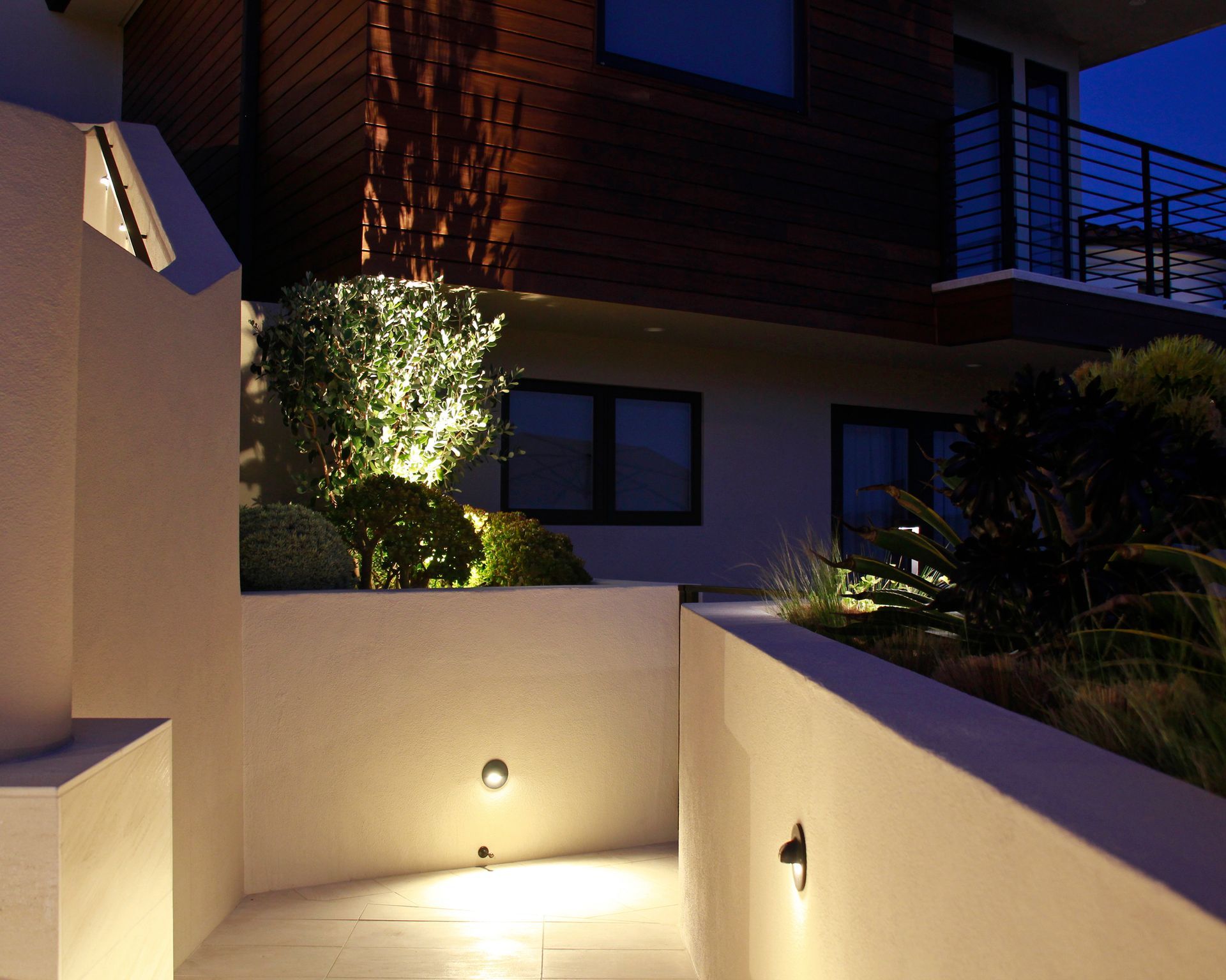 Modern apartment exterior at dusk with warm-lit walkway and landscaped shrubs