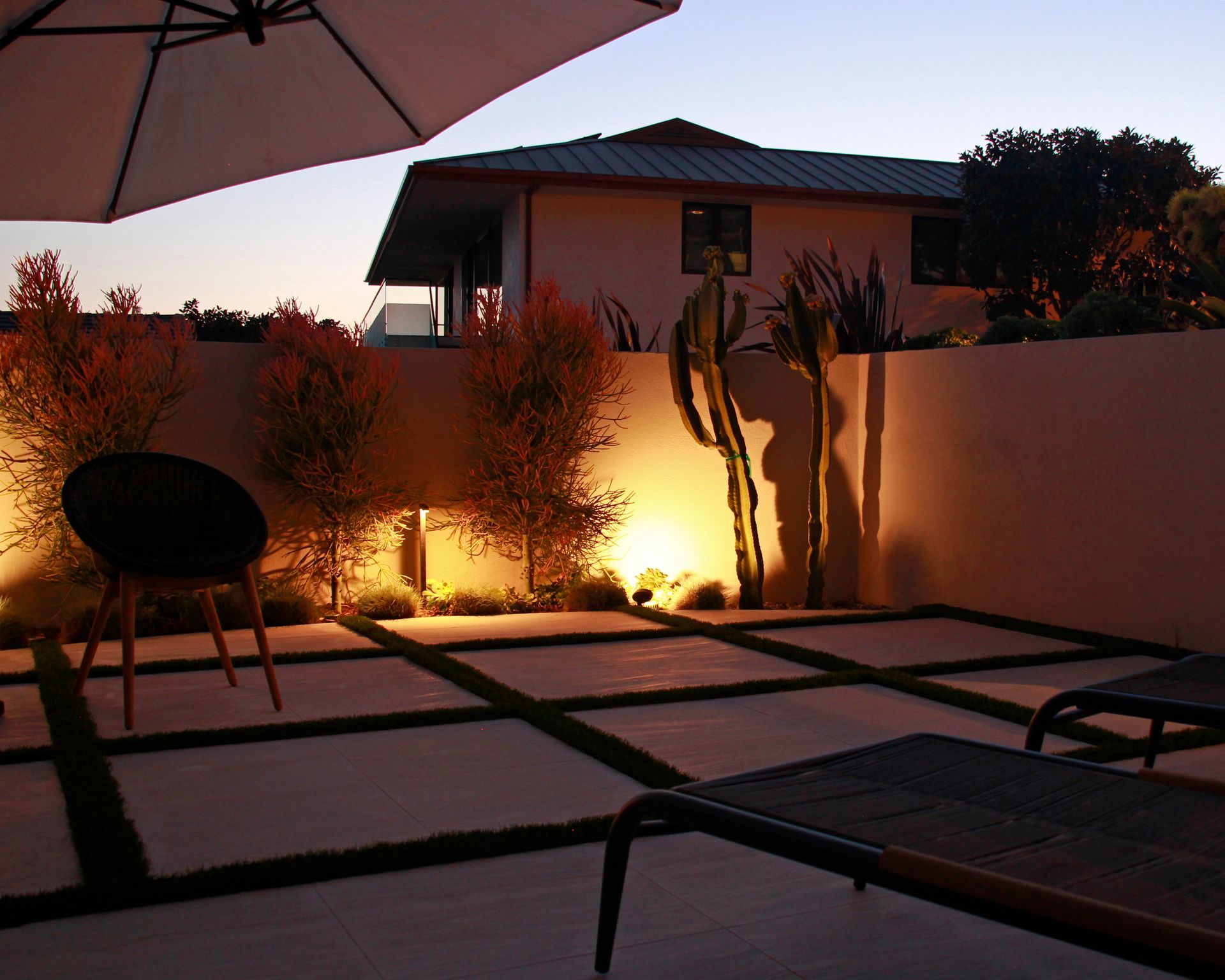 Backyard patio at dusk with lounge chairs, umbrella, and warm garden lighting against a white wall