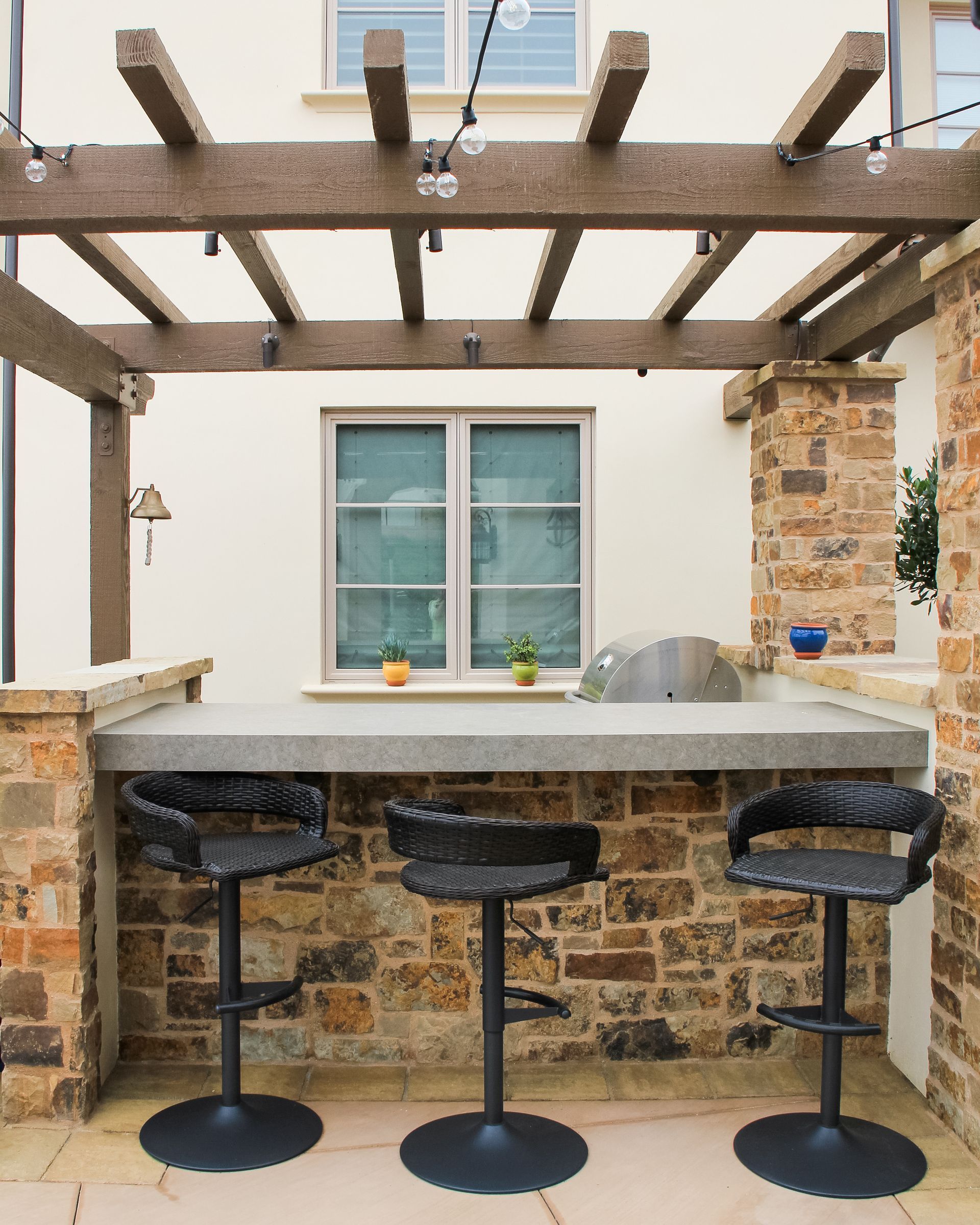 Outdoor stone bar with three stools under a pergola, in front of a window.