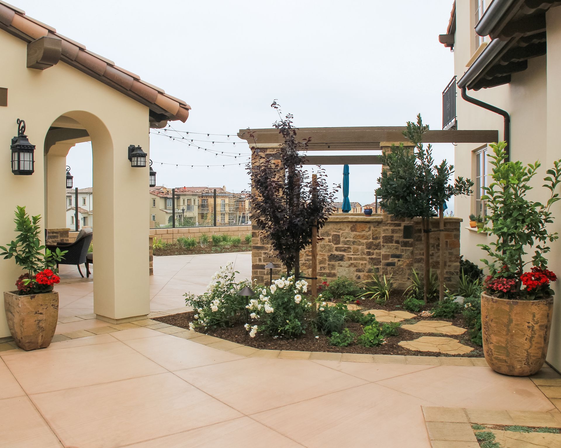 Courtyard with stone planters, potted flowers, arched entryway, and tan tiled patio between stucco buildings