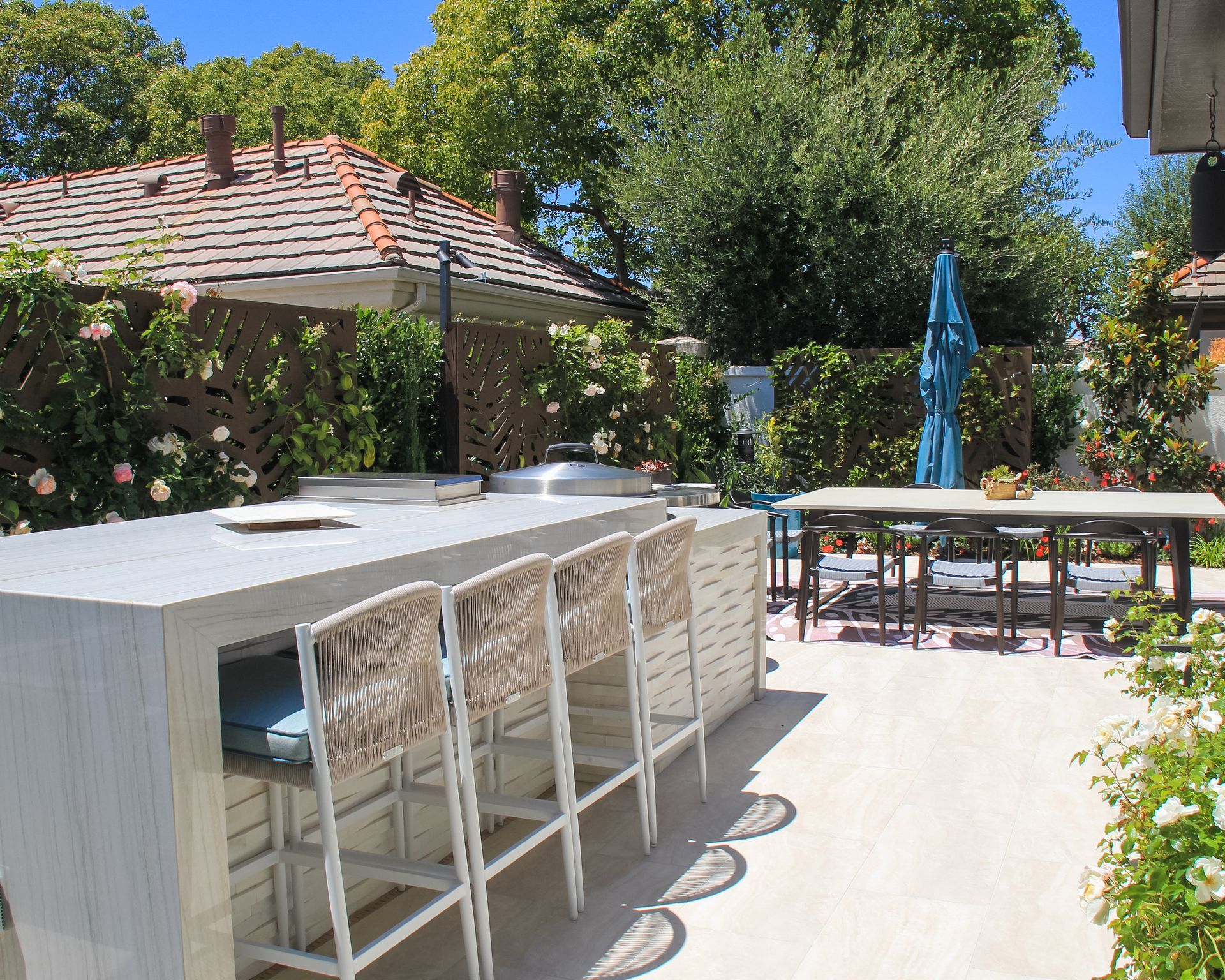 Backyard patio with white bar, wicker stools, dining table, and blue umbrella in sunlight