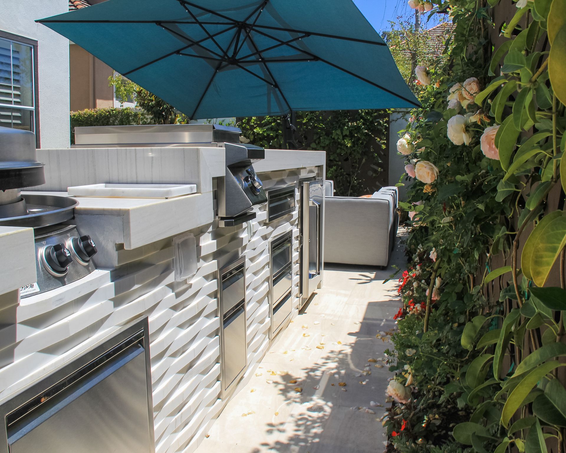 Outdoor patio with built-in grill and storage cabinets under a blue umbrella, bordered by lush greenery