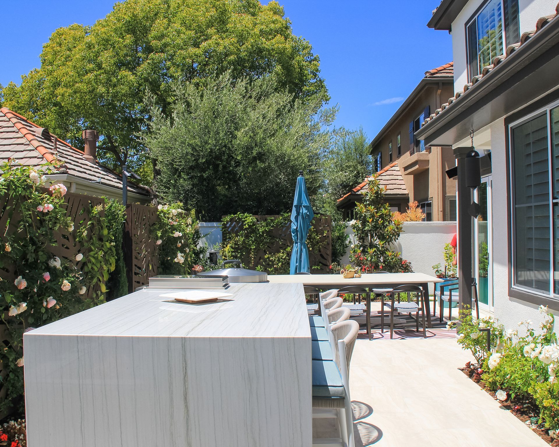 Sunny backyard patio with white outdoor dining table, blue umbrella, and lush greenery beside a house