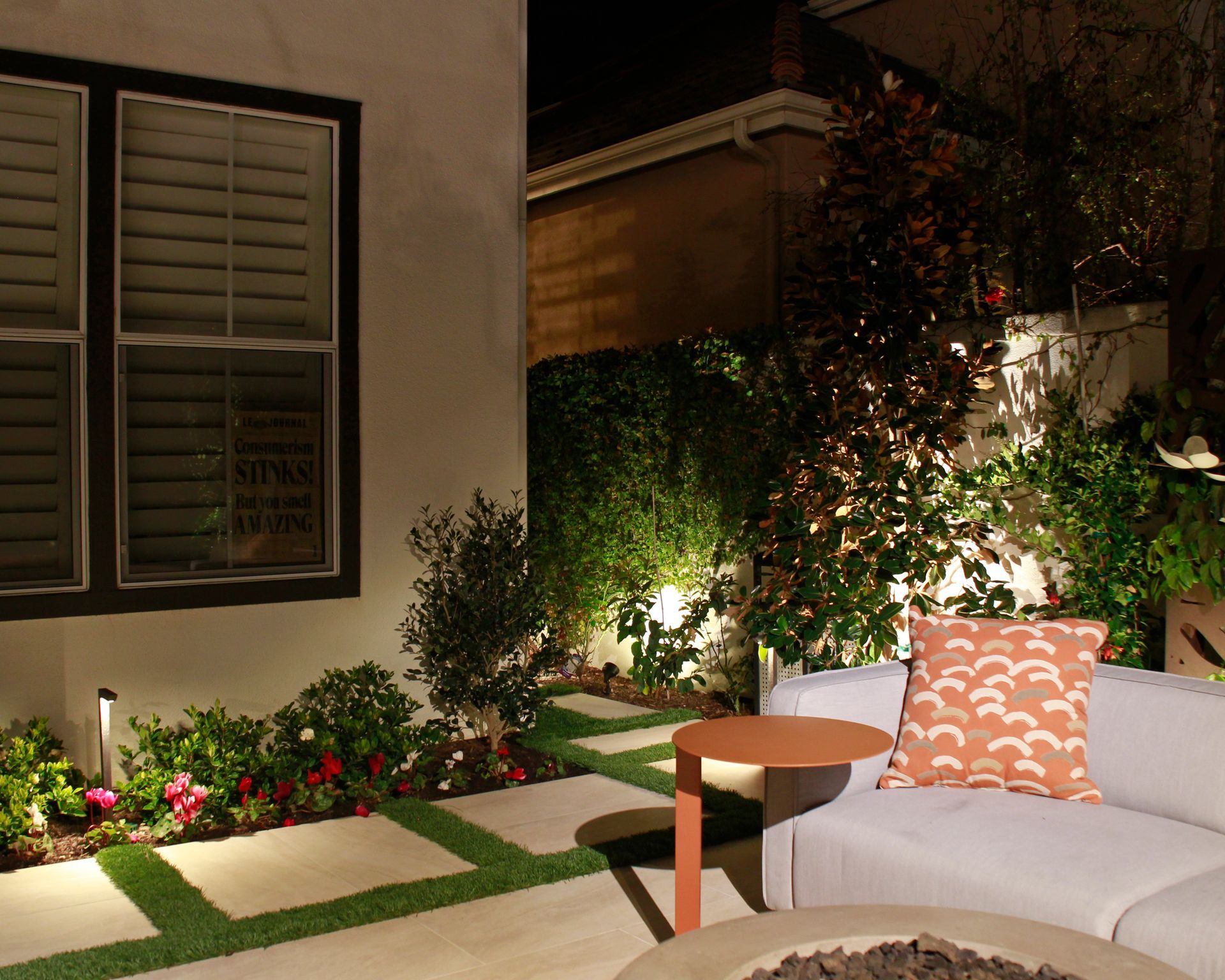 Nighttime patio with white sofa, orange pillow, potted plants, and garden lighting beside a house.