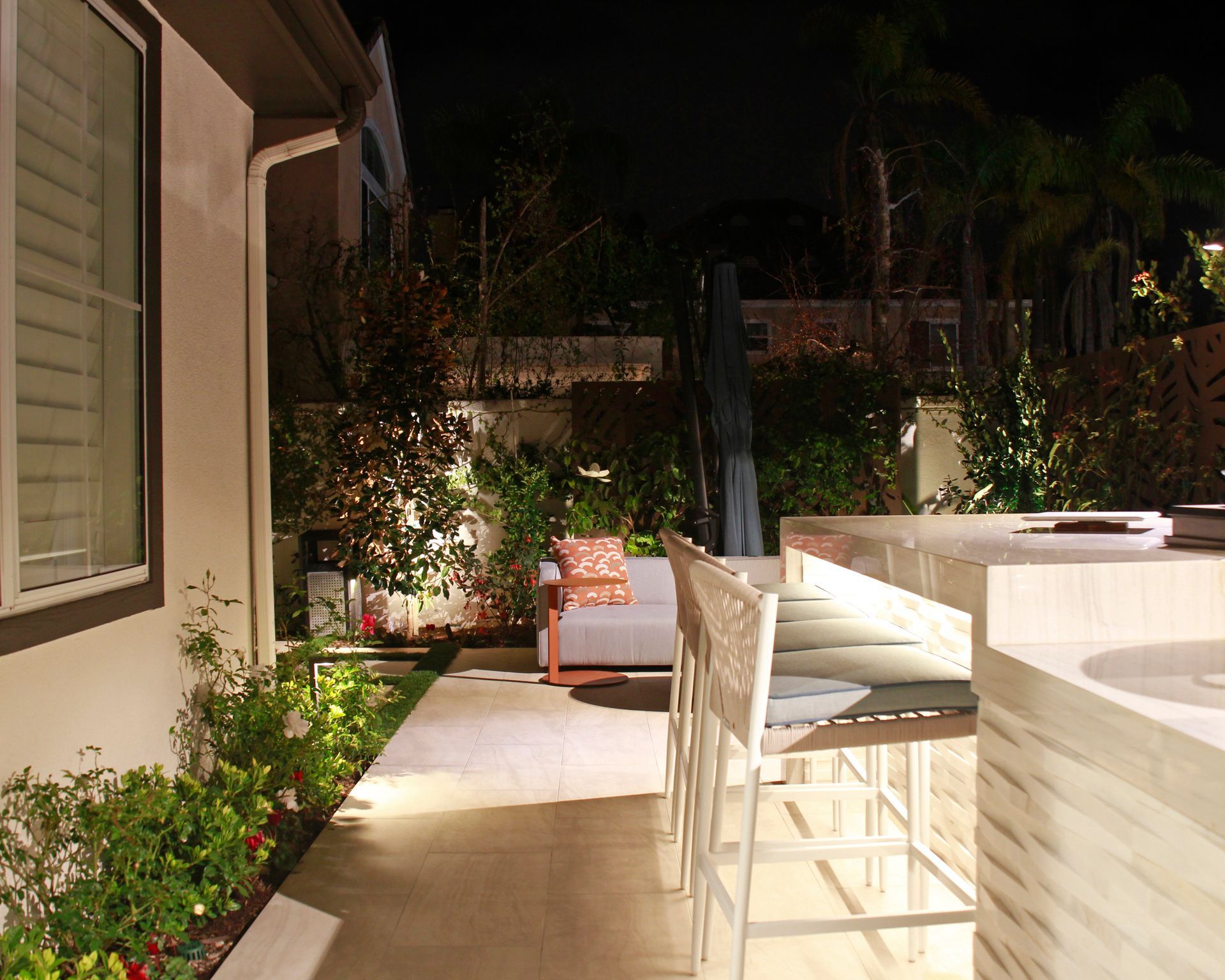 Nighttime patio with white bar stools, lit countertop, and a lounge chair beside a garden path.
