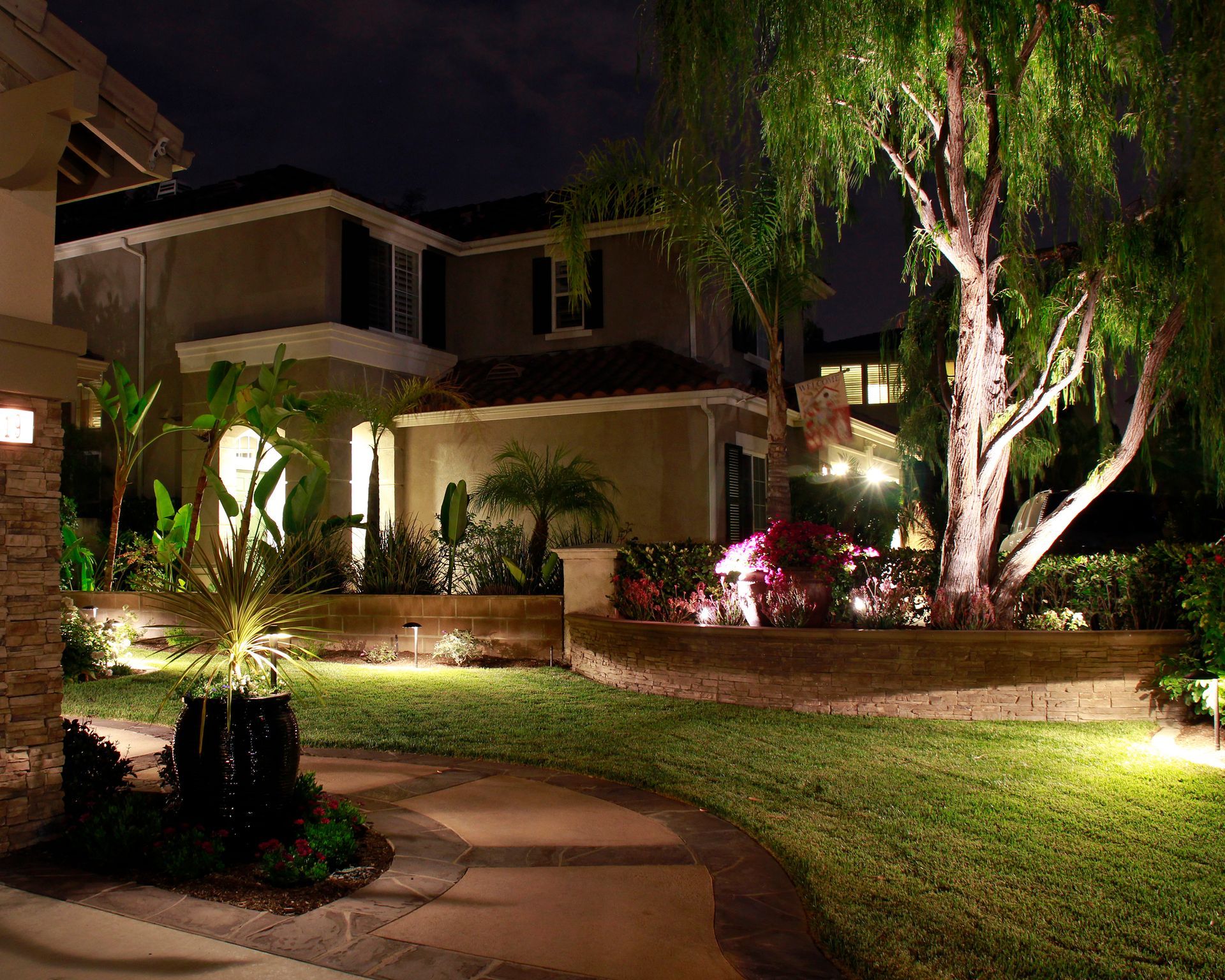 Nighttime landscaped courtyard with lit pathways, a large tree, and apartment homes in the background