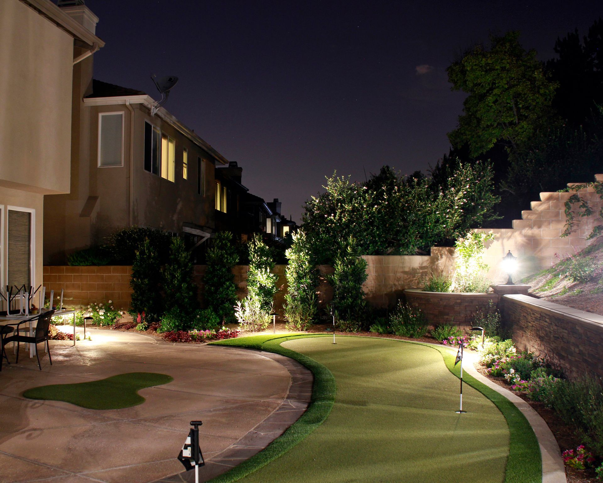 Nighttime backyard with lit curved lawn, patio, and landscaped garden beside a house