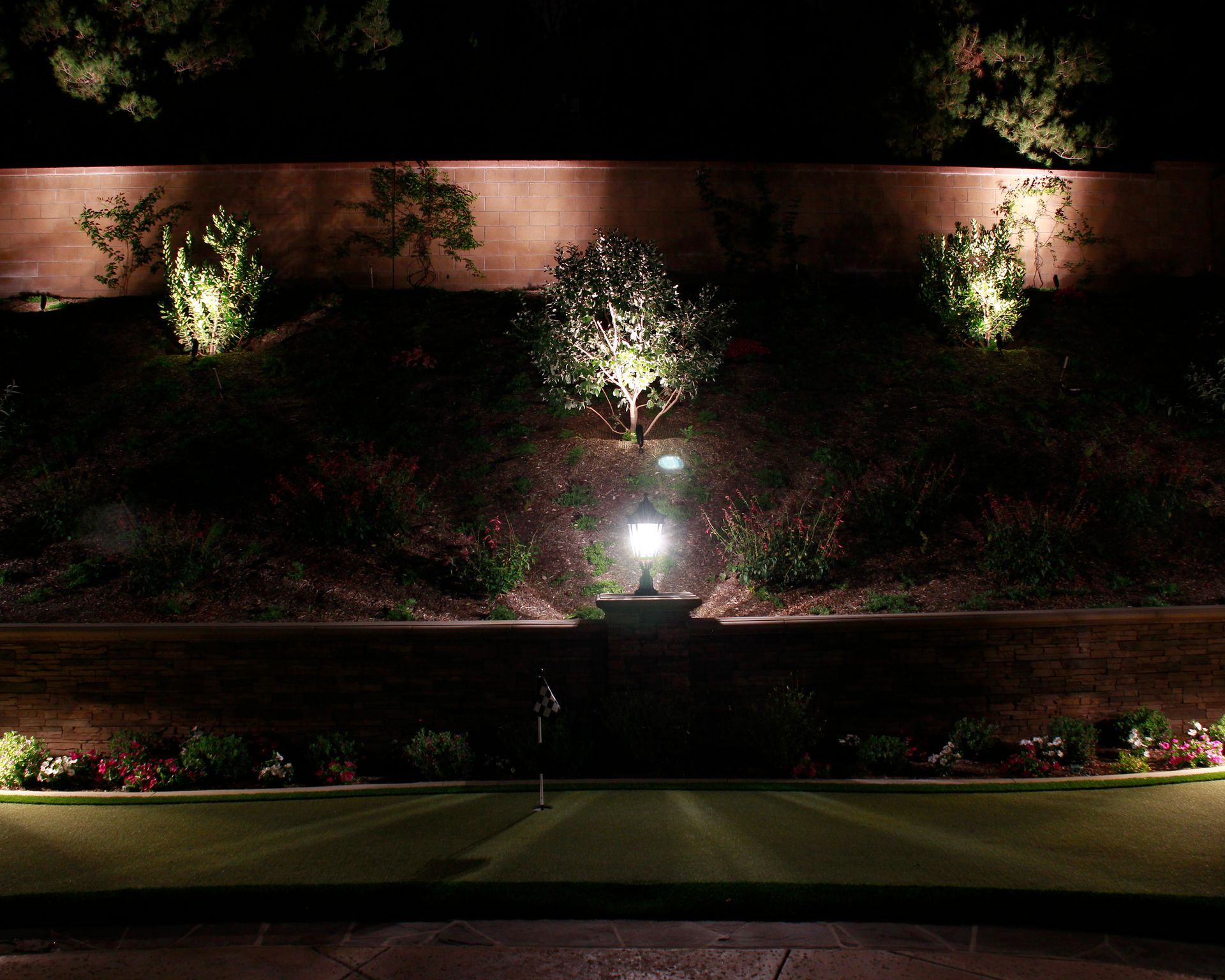 Nighttime landscaped walkway lit by ground lights along a wall and trees