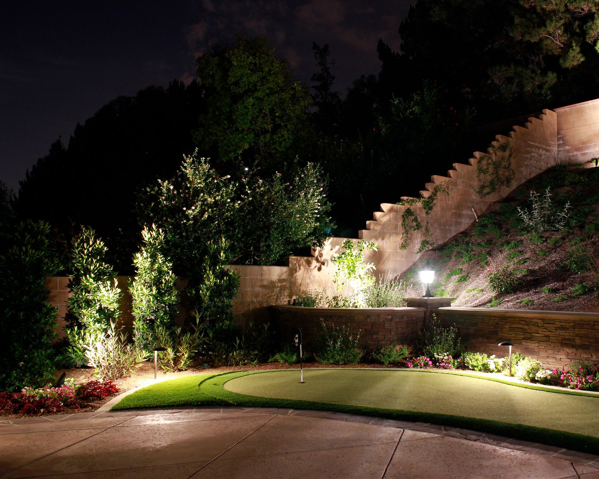 Lit landscaped garden at night with trees, stone walls, and a curved driveway