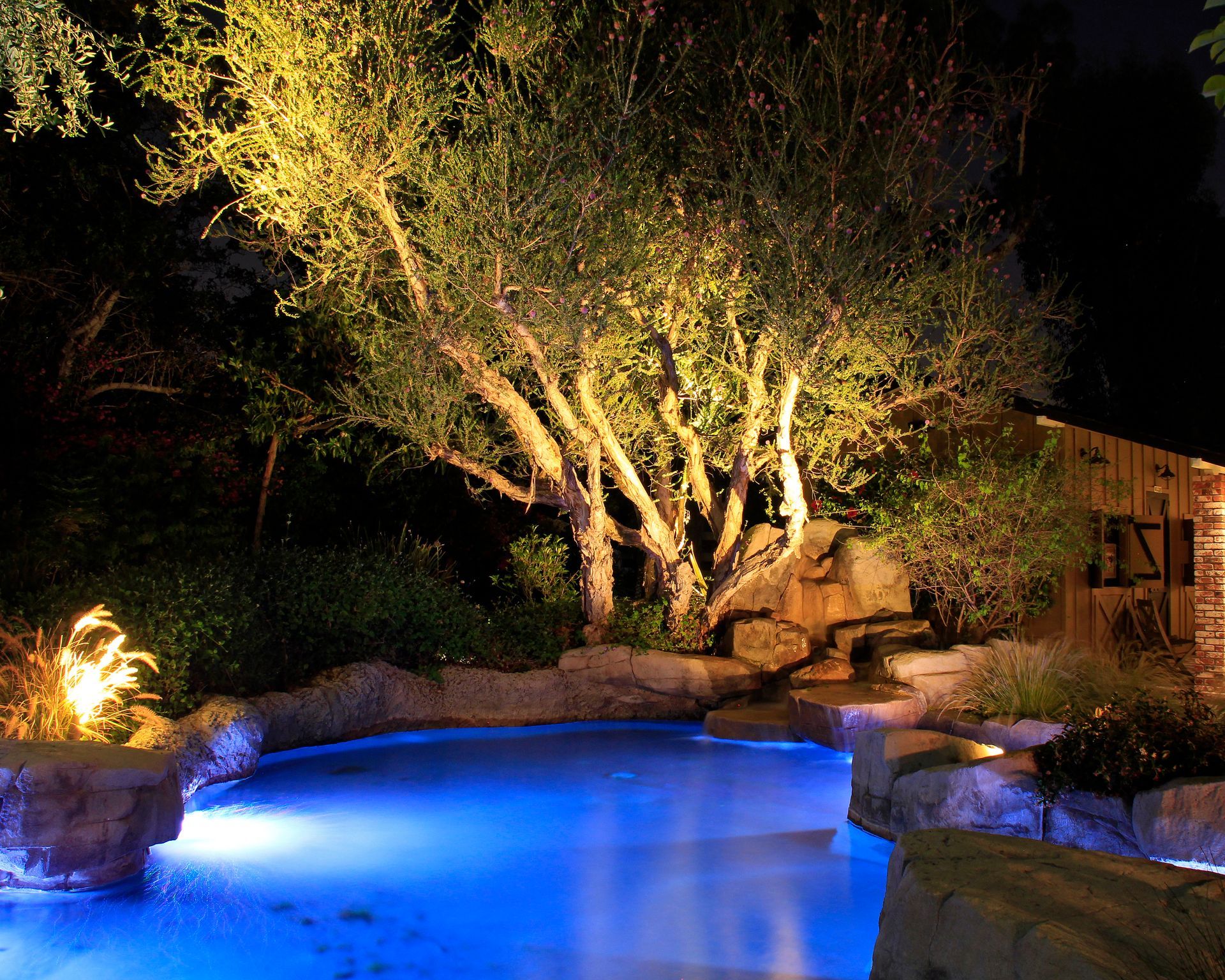 Illuminated backyard pool at night with blue water, stone edges, and trees lit by warm landscape lighting