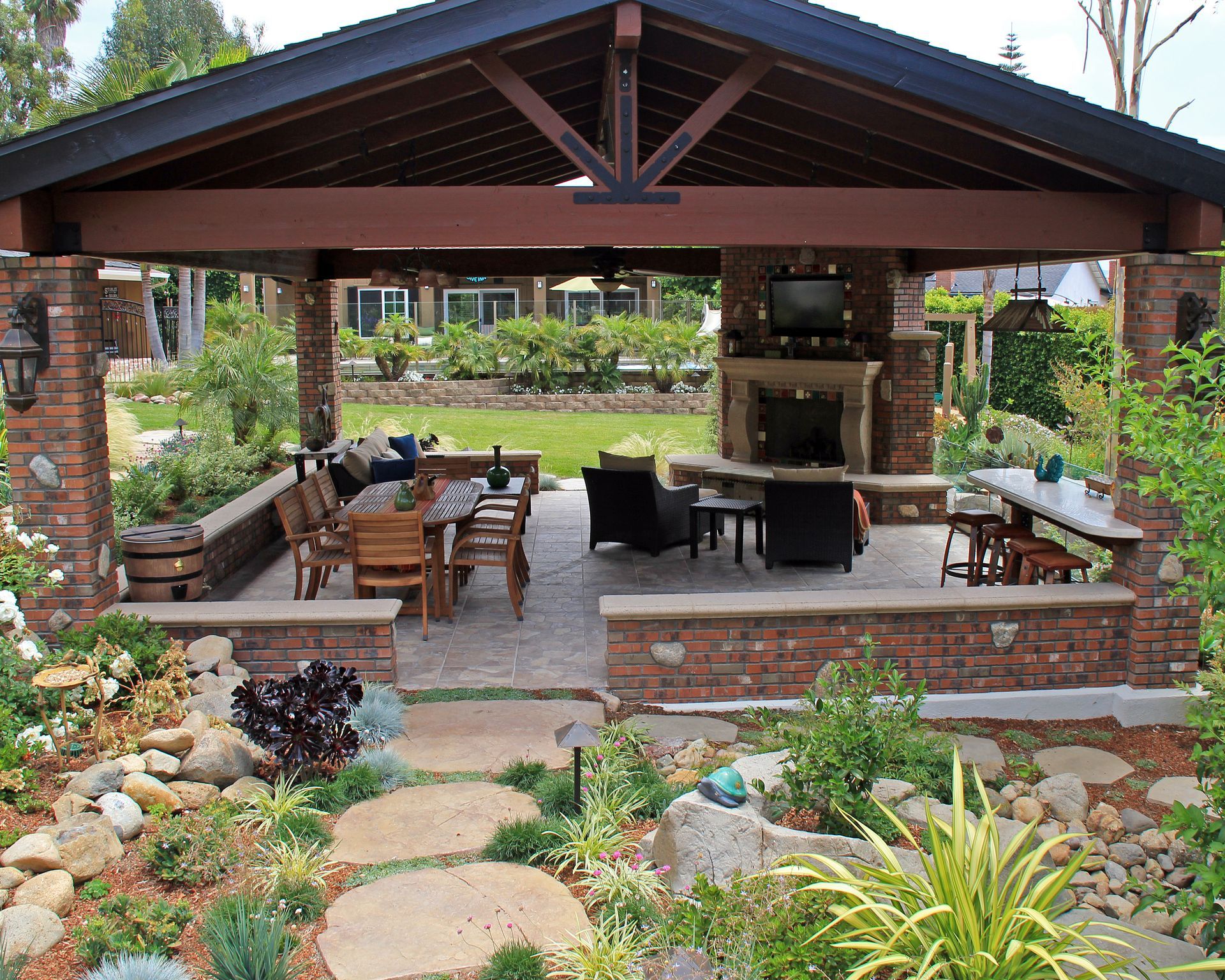 Covered outdoor patio with brick seating, wooden beams, and lush garden landscaping around stepping stones