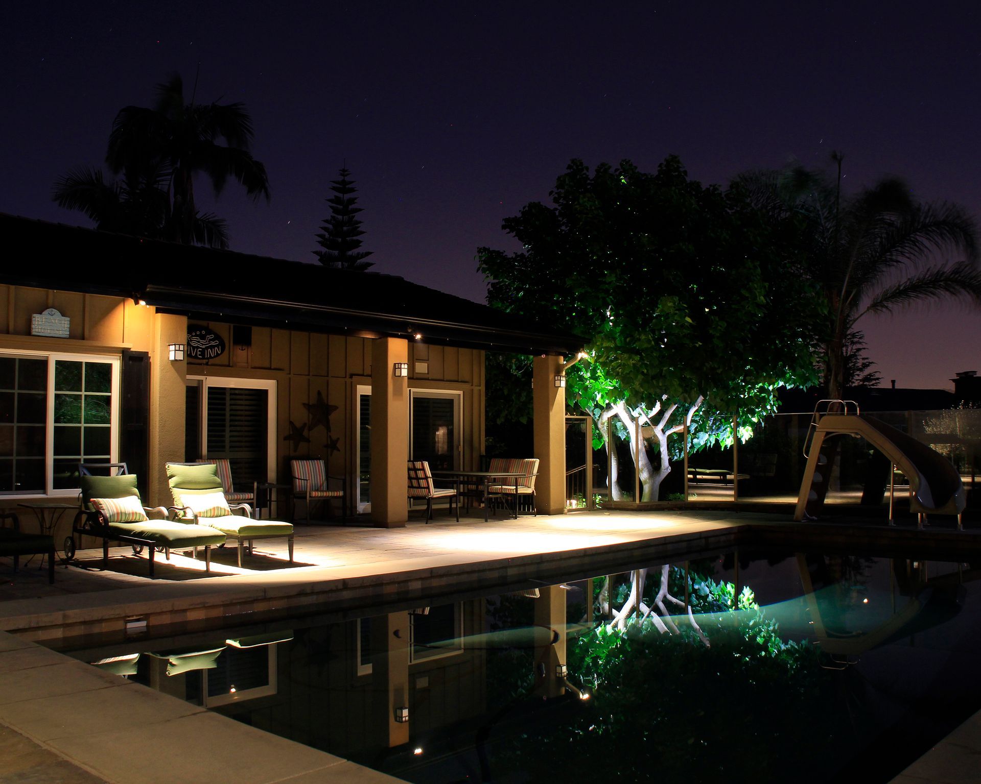 Nighttime patio with lounge chairs, lit trees, and a pool reflecting the house lights