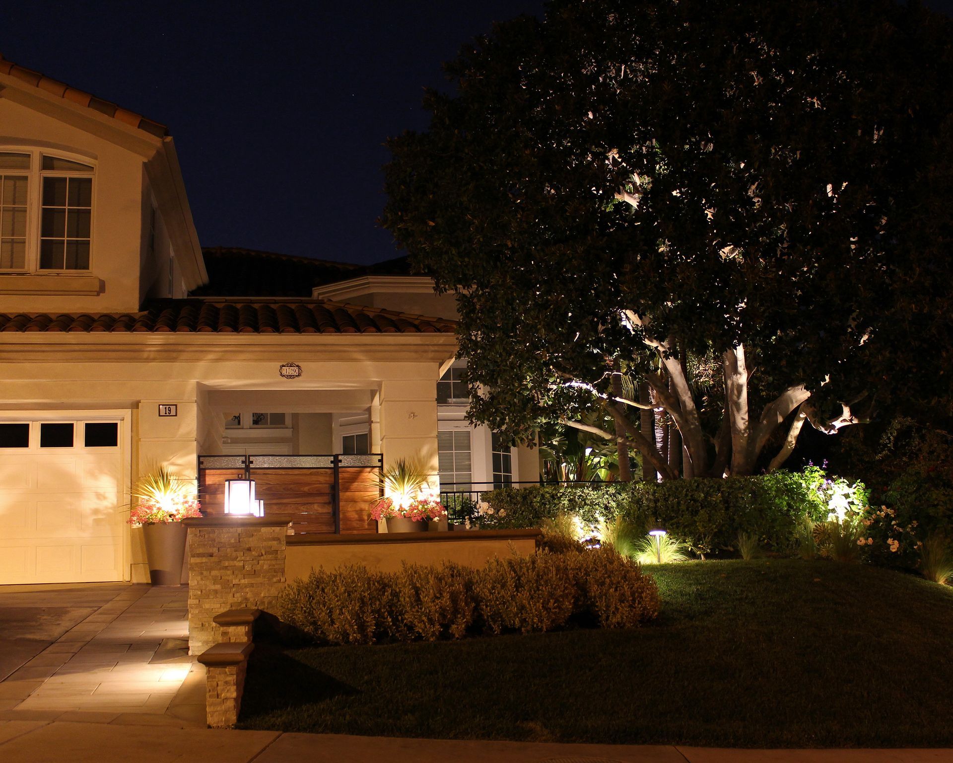Nighttime view of a lit suburban house with a large tree and landscaped front yard