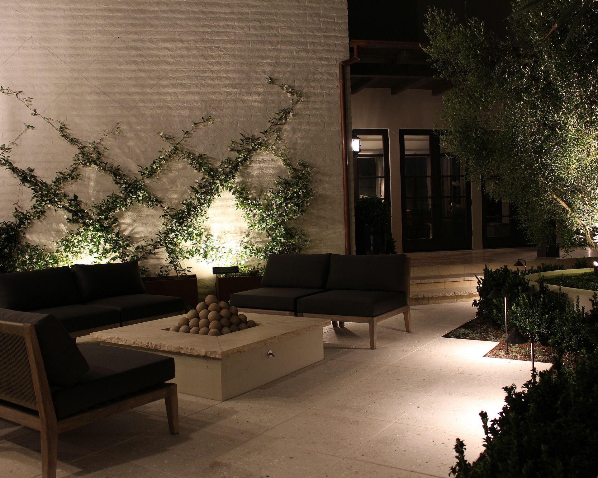 Modern outdoor patio at night with lounge chairs, low table, and lit trellis vines beside a building