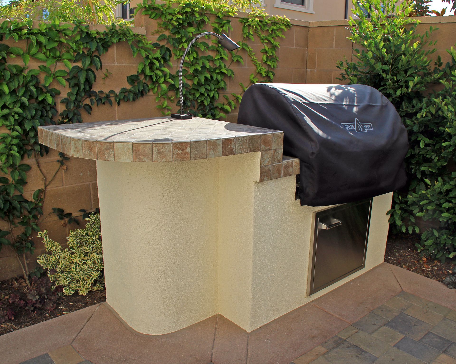 Outdoor grill beside a tan stucco counter and brick patio, with green vines along the wall