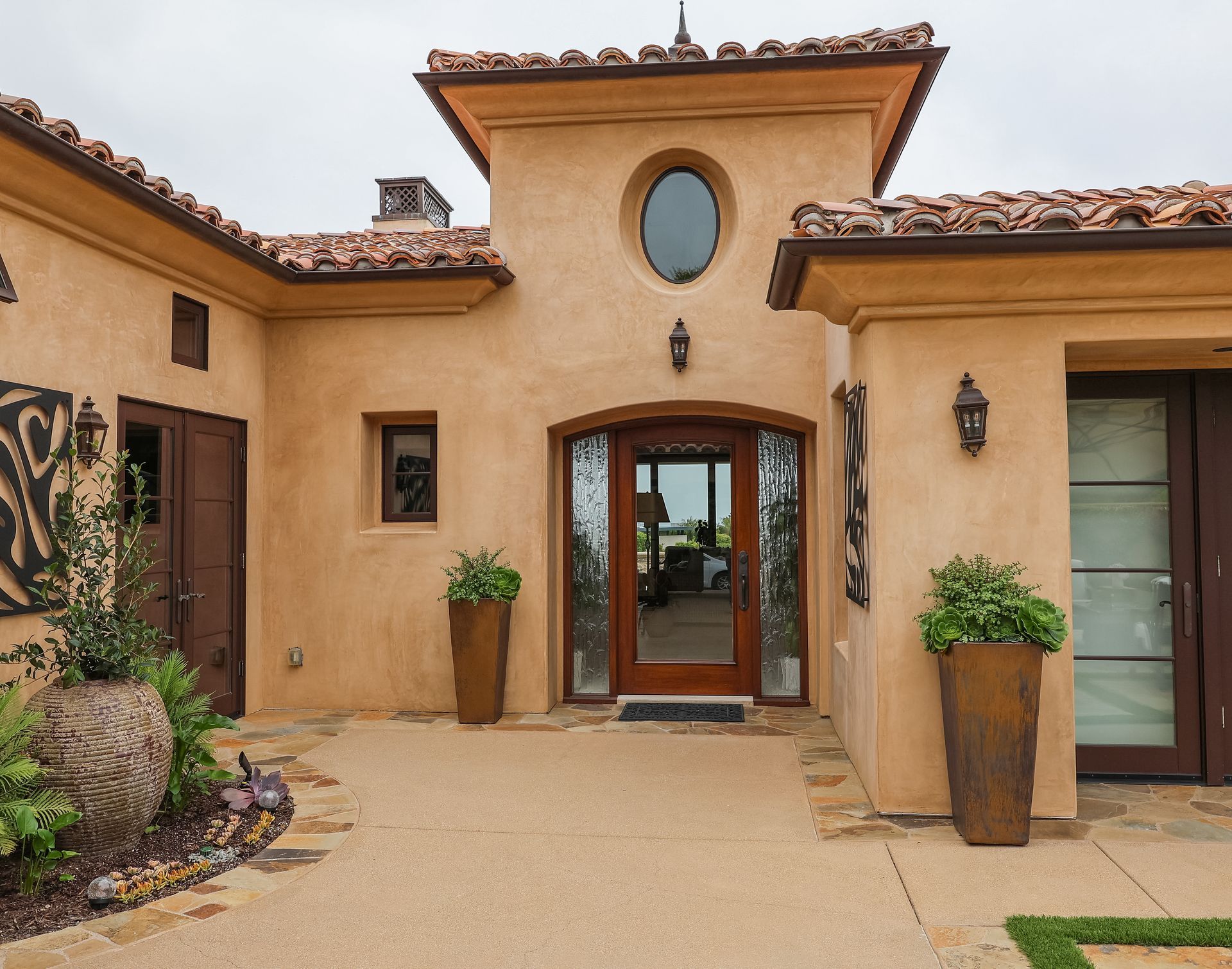 Stucco house entrance with wooden door, oval window, potted plants, and terracotta roof tiles