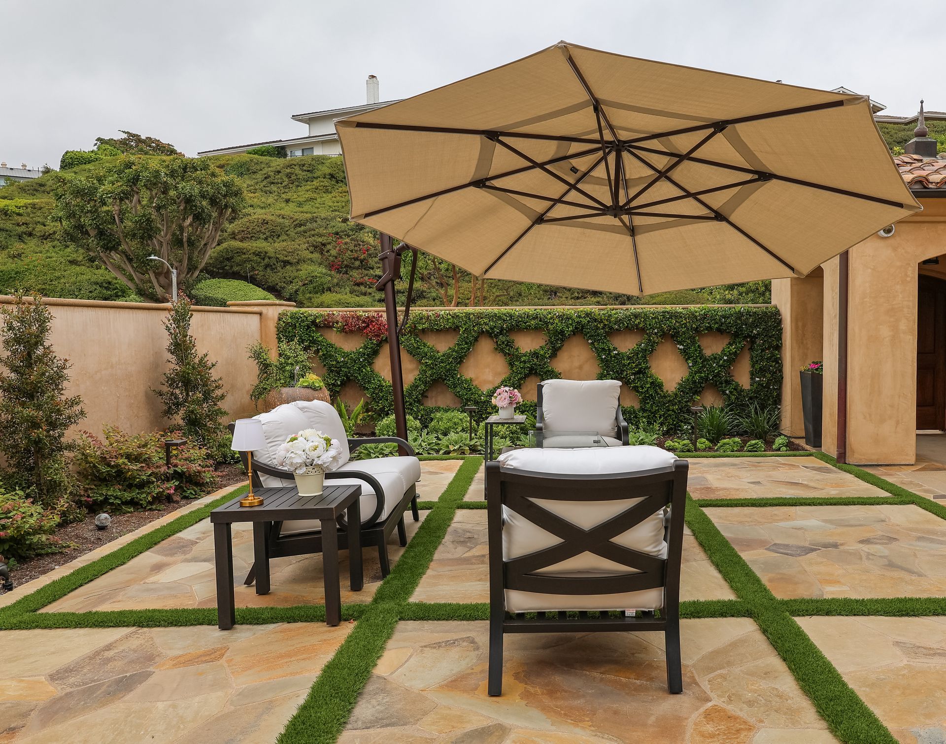 Outdoor patio with tan umbrella, lounge chairs, and greenery along a stone path and wall