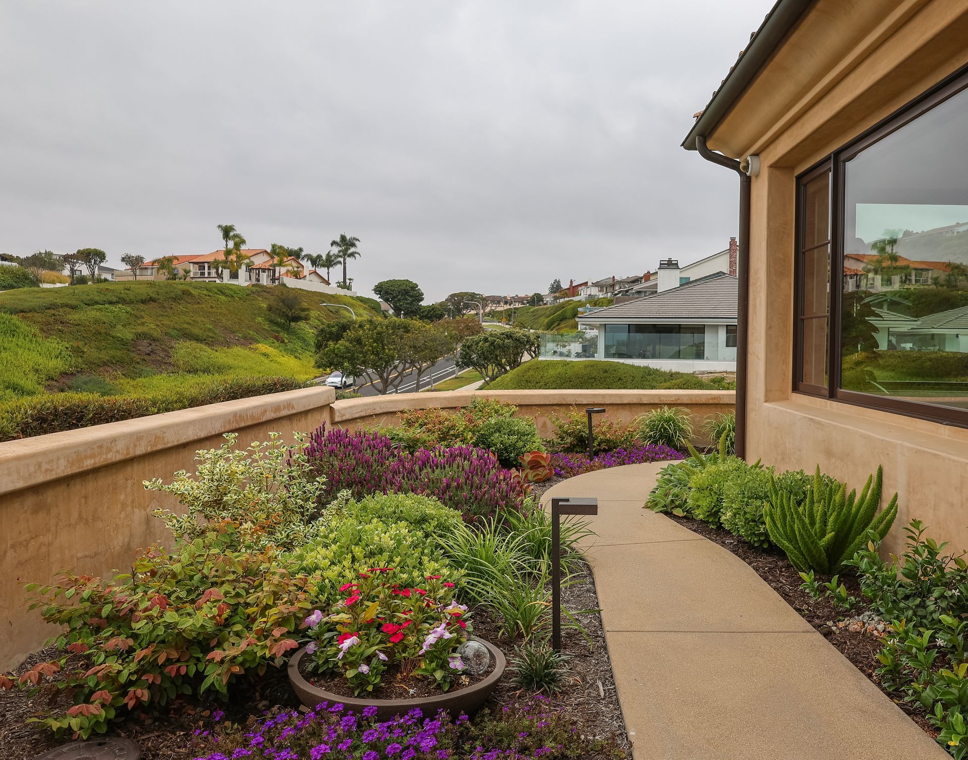 Courtyard garden path beside a tan house, with colorful flowers and a cloudy coastal view.