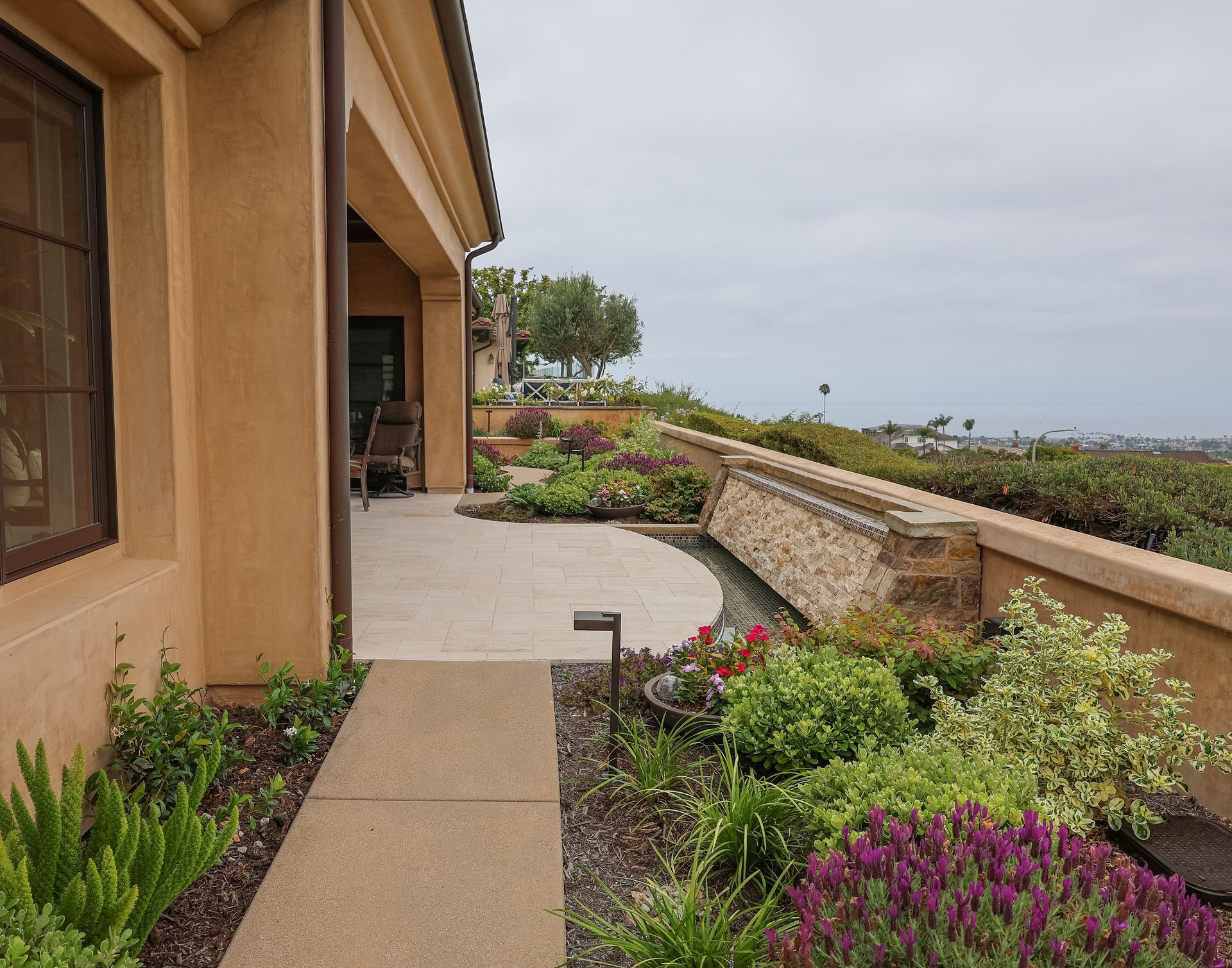 Stucco patio walkway beside a garden and low wall overlooking a coastal view