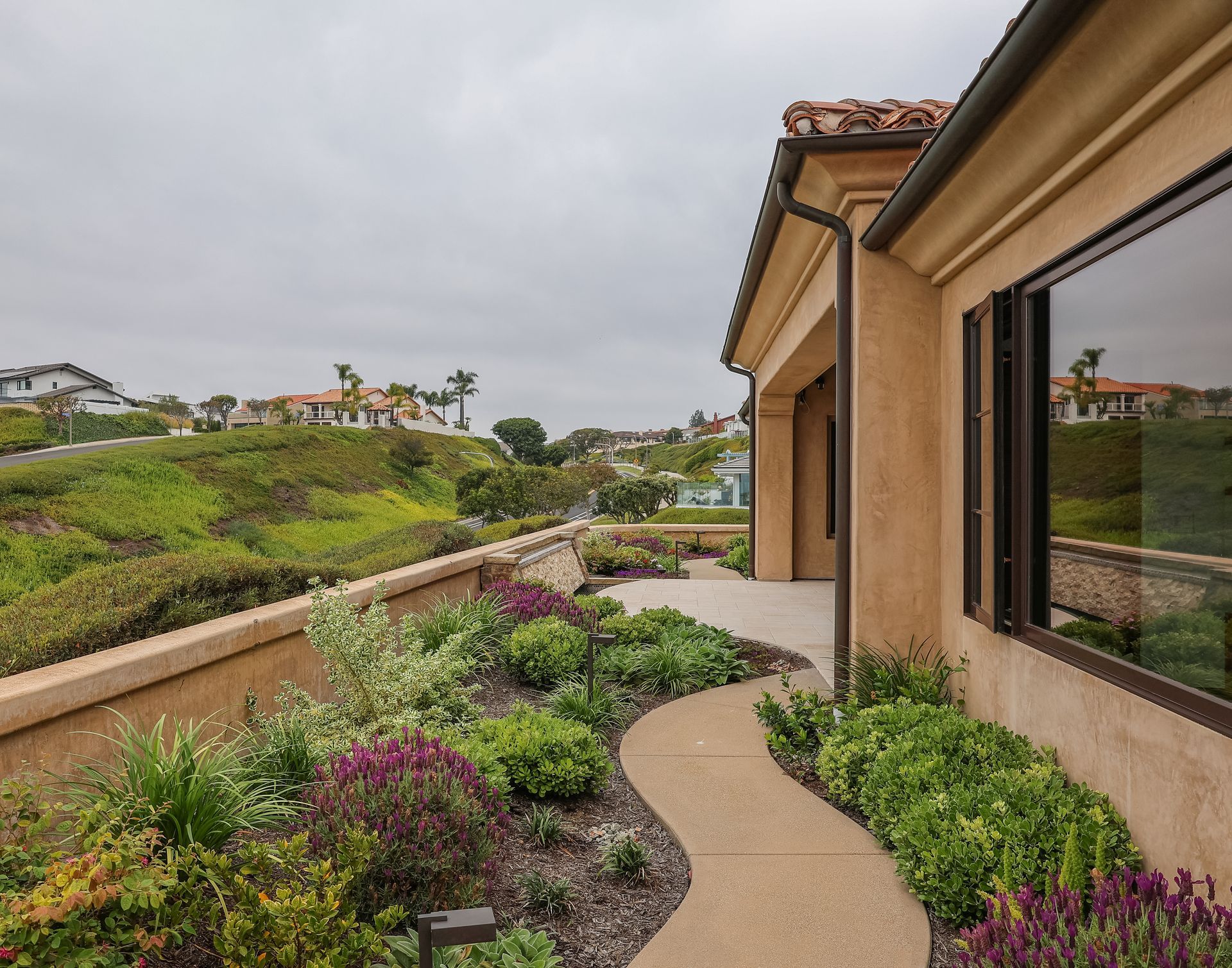 Curved garden path beside a tan house, bordered by flowering shrubs and overlooking a green hillside.