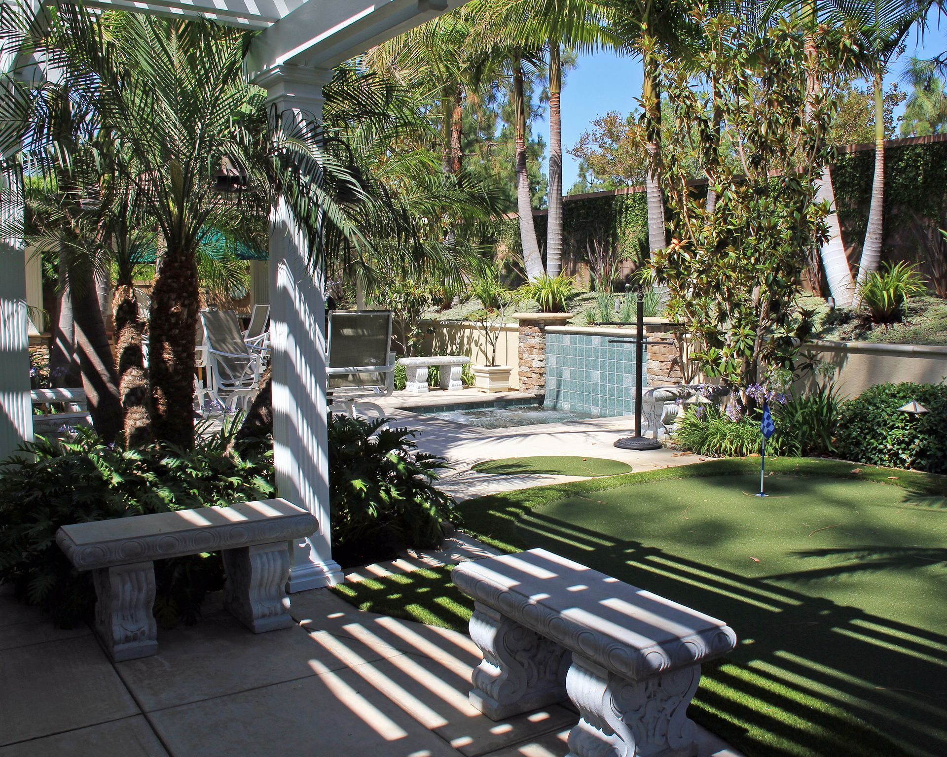 Shaded patio with stone benches, artificial grass, palm trees, and a small pool fountain.