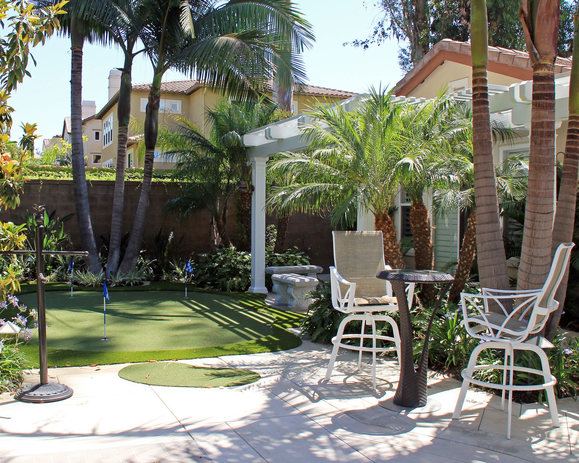 Backyard patio with palm trees, lawn, and white outdoor chairs beside a brick wall and pergola.