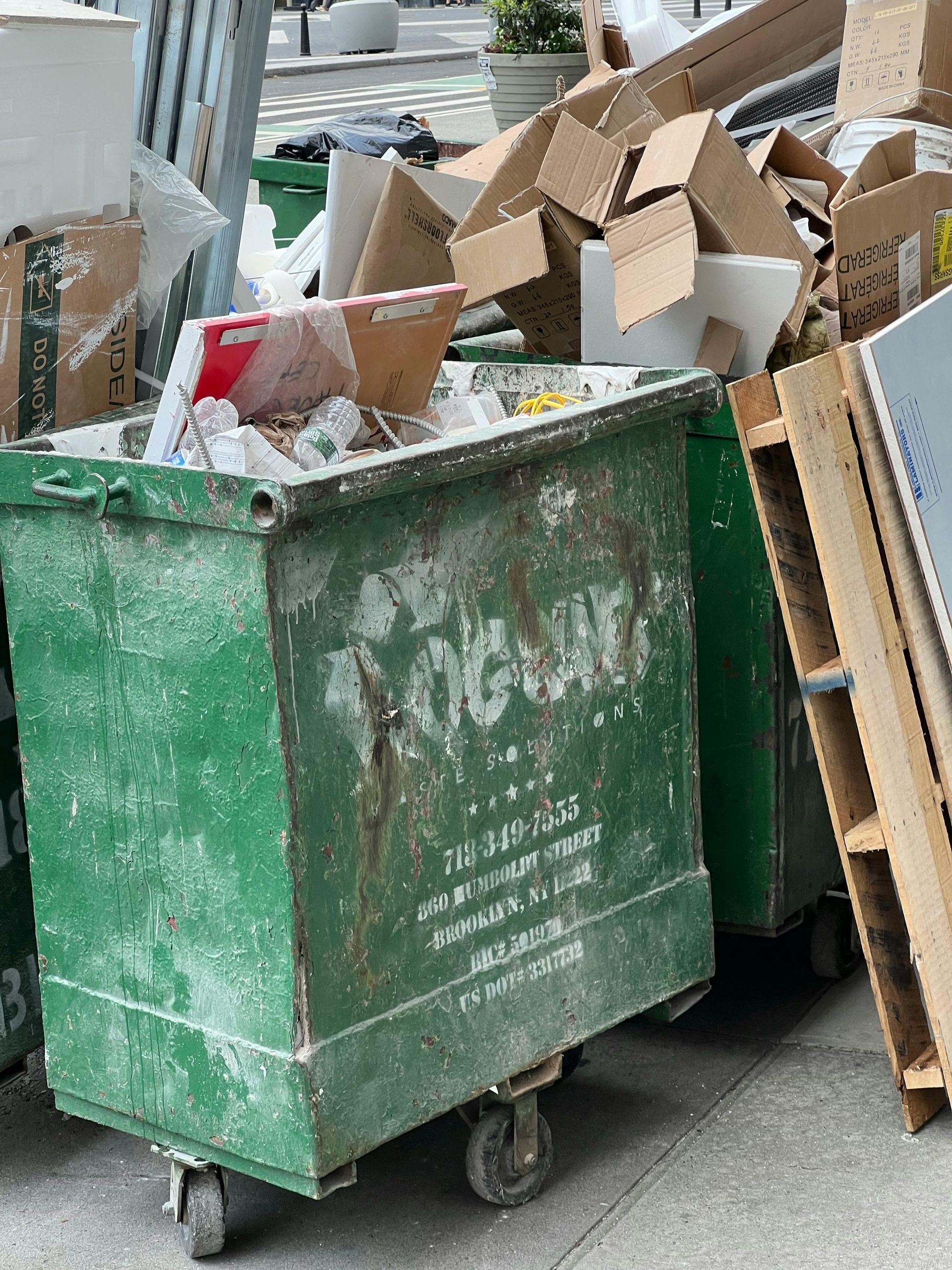 A green, weathered industrial dumpster overflowing with cardboard boxes and debris on a city sidewalk.