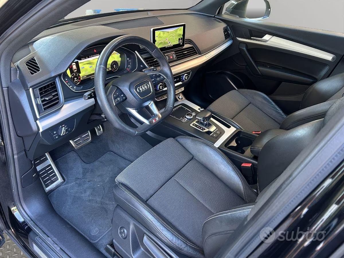 Interni dell'auto con sedili trapuntati neri, volante, schermo del cruscotto e console centrale illuminati alla luce del giorno.