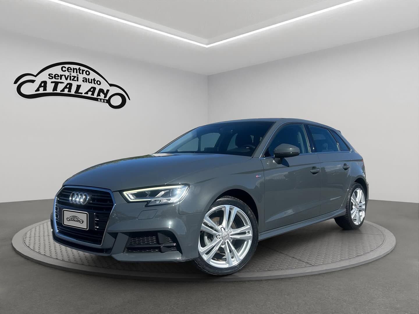 Una berlina Audi grigia su una piattaforma espositiva girevole in uno showroom bianco con l'insegna Catalano.