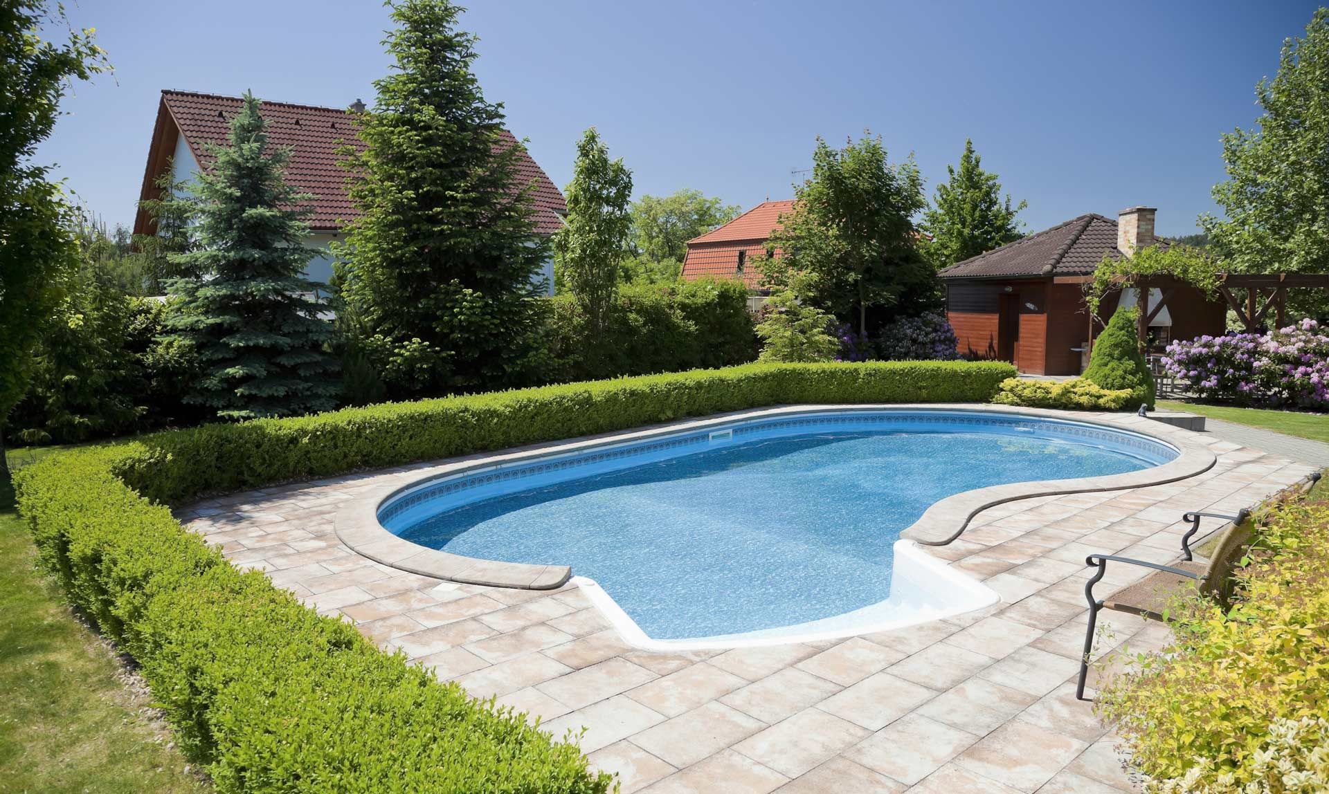 Pool with clear blue water surrounded by stone patio, green hedges, and trees.