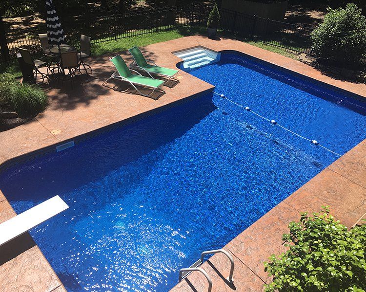 Blue rectangular swimming pool with diving board, surrounded by a brown patio, lounge chairs, and an outdoor dining set.